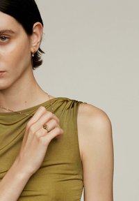 Top sin mangas de color verde oliva con un diseño drapeado, acompañado de un delicado collar de capas y un anillo de oro con un diseño circular.