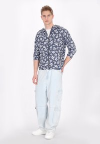 Jachetă cu fermoar, cu glugă, în nuanțe florale de navy, purtată cu pantaloni cargo de un albastru deschis. Ținuta are o croială lejeră și este completată cu adidași albi.