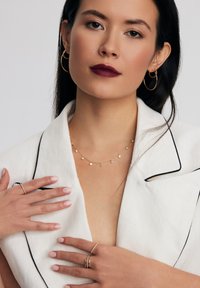 Złote kolczyki w kształcie kół, delikatny złoty naszyjnik z małymi okrągłymi akcentami, biały żakiet z czarnym obszyciem i zadbane paznokcie w odcieniu nude.