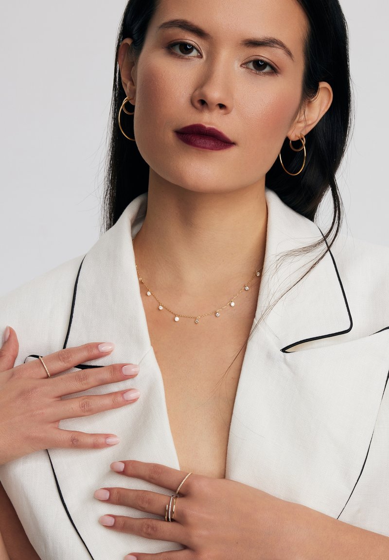 Złote kolczyki w kształcie kół, delikatny złoty naszyjnik z małymi okrągłymi akcentami, biały żakiet z czarnym obszyciem i zadbane paznokcie w odcieniu nude.