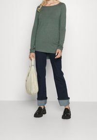 Grünes Langarmshirt mit U-Ausschnitt, kombiniert mit dunkelblauer Jeans, die am Saum hochgekrempelt ist, und schwarzen Plateau-Schuhen. Es wird eine leichte Tasche gehalten.