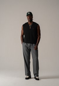 Schwarzer ärmelloser Pullover, graue maßgeschneiderte Hose, schwarze Schuhe mit Netzdetails und eine grüne Baseballmütze mit weißem Logo. Sichtbare goldene Schmuckdetails.