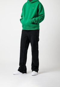 Persona che indossa un hoodie verde, pantaloni cargo neri e sneakers bianche in piedi davanti a uno sfondo bianco semplice.