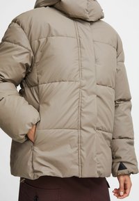 Polstret jakke i taupe med hætte, med et quiltet design, elastiske manchetter, sidelommer og en lille sort logoaccent.