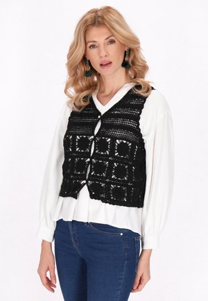 Lyshåret kvinde iført grønne frynseøreringe, hvid bluse med lange ærmer, sort hæklevest og blå jeans, stående mod hvid baggrund.