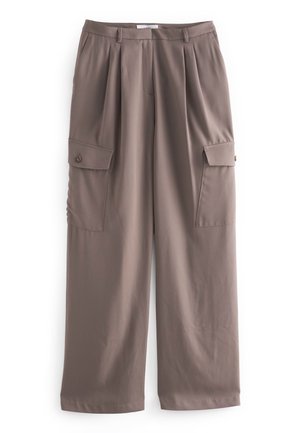 Kvinde står med armene krydsede iført beige kortærmet top, taupe vid, bredbenede cargobukser og cremefarvede sandaler med stropper i et lyst rum.