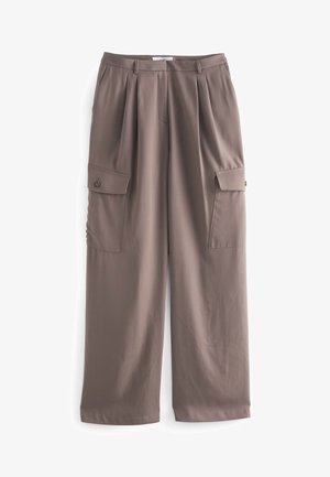 Vrouw staat met armen over elkaar, draagt beige top met korte mouwen, taupekleurige wijde cargobroek en crèmekleurige sandalen met bandjes in een lichte kamer.