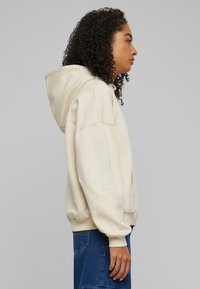 Beige hettegenser laget av myk stoff, med avslappet passform, ribbede mansjetter og en frontlomme. Stilt med blå denim.