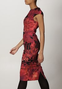 Robe rouge à motif floral avec une silhouette ajustée, des manches courtes, et un ourlet à hauteur des genoux, présentant une fente latérale et des accents noirs.