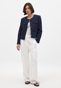 Tamsiai mėlyna tekstūruota striukė su apvaliu apykakle, dviem priekinėmis kišenių ir keturiais mygtukais, dėvima ant baltos palaidinės ir derinama su baltomis plačiomis kelnėmis.