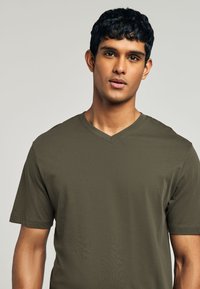 Jonge man met kort zwart haar, die een olijfgroen T-shirt met V-hals en korte mouwen draagt, kijkt recht vooruit tegen een effen lichte achtergrond.