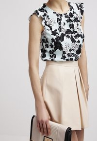 Blusa senza maniche floreale con fiori in bianco e nero abbinata a una gonna beige a vita alta con pieghe, mentre tiene una borsa beige e nera.