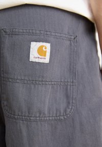 Grå fishtail-stoff med en baklomme som har et hvitt merke og en gul logo. Sømmen er synlig langs kantene.
