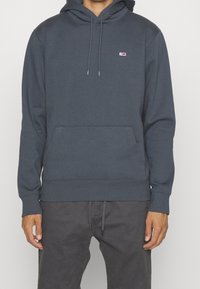 Persona con una sudadera con capucha gris oscuro con bolsillo frontal y un pequeño logo rectangular en el pecho, combinada con pantalones gris oscuro.