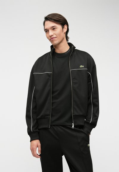 Veste de survêtement noire avec des détails en blanc, fermeture éclair, poignets côtelés et logo Lacoste sur la poitrine. Associée à un haut noir assorti.
