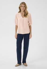 Blusa rosa chiaro con scollo a V e maniche corte a sbuffo, abbinata a jeans dritti blu scuro e ballerine beige.