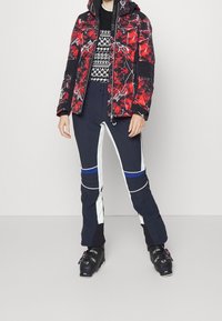 Röd och svart mönstrad puffjacka med huva över en svart mönstrad tröja. Marinblå skidbyxor med vita och blå detaljer. Svarta skidstövlar.