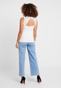 Kvinna med lockigt hår iklädd vit topp med spetsbak, ljusblå vida jeans och svarta högklackade skor, vänd bort.