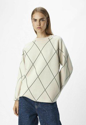 Suéter beige con patrón de diamantes negros, cuello redondo y mangas holgadas, combinado con jeans azules texturizados sobre un fondo liso.