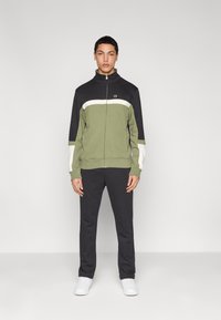 Veste de survêtement zippée couleur vert olive avec un effet bloc de couleurs noir et blanc, assortie à un pantalon noir et des baskets blanches. Texture de tissu lisse.
