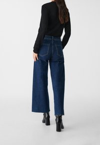 High-waisted, wide-leg blue jeans with a raw hem and two back pockets, paired with a black long-sleeve top and black heeled boots.