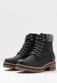 Bottes noires à lacets montantes avec semelles épaisses marron clair et noires, poignets en tricot gris rembourrés et matériau texturé semblable au cuir.