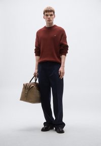 Πλεκτό μπεζ πουλόβερ με ρίγες, ναυτικό παντελόνι φαρδύ, καφέ δερμάτινη τσάντα. Το μοντέλο στέκεται με τα χέρια στα πλάγια, μαύρα παπούτσια, ουδέτερο φόντο.