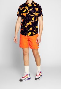 Mann mit schwarzem kurzärmligen Hemd mit orangefarbenem abstraktem Muster, leuchtend orangefarbigen Shorts, weißen Socken und mehrfarbigen Sportschuhen.