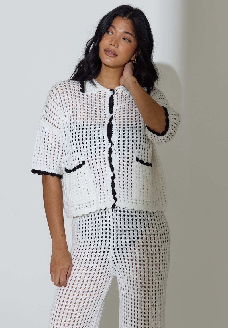 Cárdigan blanco de punto de crochet con mangas cortas, bordes ondulados, ribete negro y pantalones a juego con patrones y detalles de textura similares.