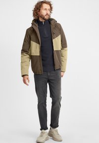 Brown and olive color-blocked jacket with a zipper, paired with a navy sweater, dark jeans, and beige shoes. Features a hood and elastic cuffs.