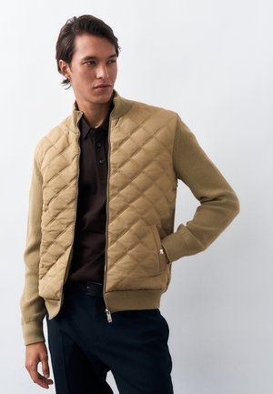 Férfi, aki bélelt, barna kabátot visel kötött ujjakkal, sötétbarna galléros póló és tengerészkék nadrág fölött, sima fehér háttér előtt áll.