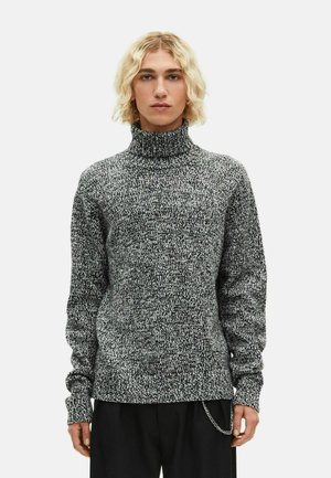 Pessoa jovem com cabelo loiro até aos ombros, vestindo um suéter de gola alta em preto e branco mesclado e calças pretas com um acessório de corrente.