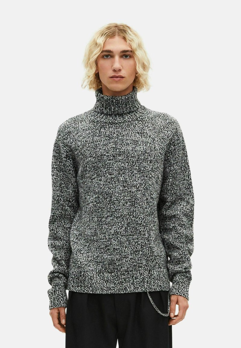 Jaunas žmogus su pečių ilgio šviesiais plaukais, vilkintis juodai baltą marmurinį golfą ir juodas kelnes su grandinės aksesuaru.