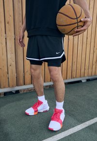 Basketball player in black shorts with white accents, wearing pink and white sneakers, holding a basketball, against a wooden backdrop.