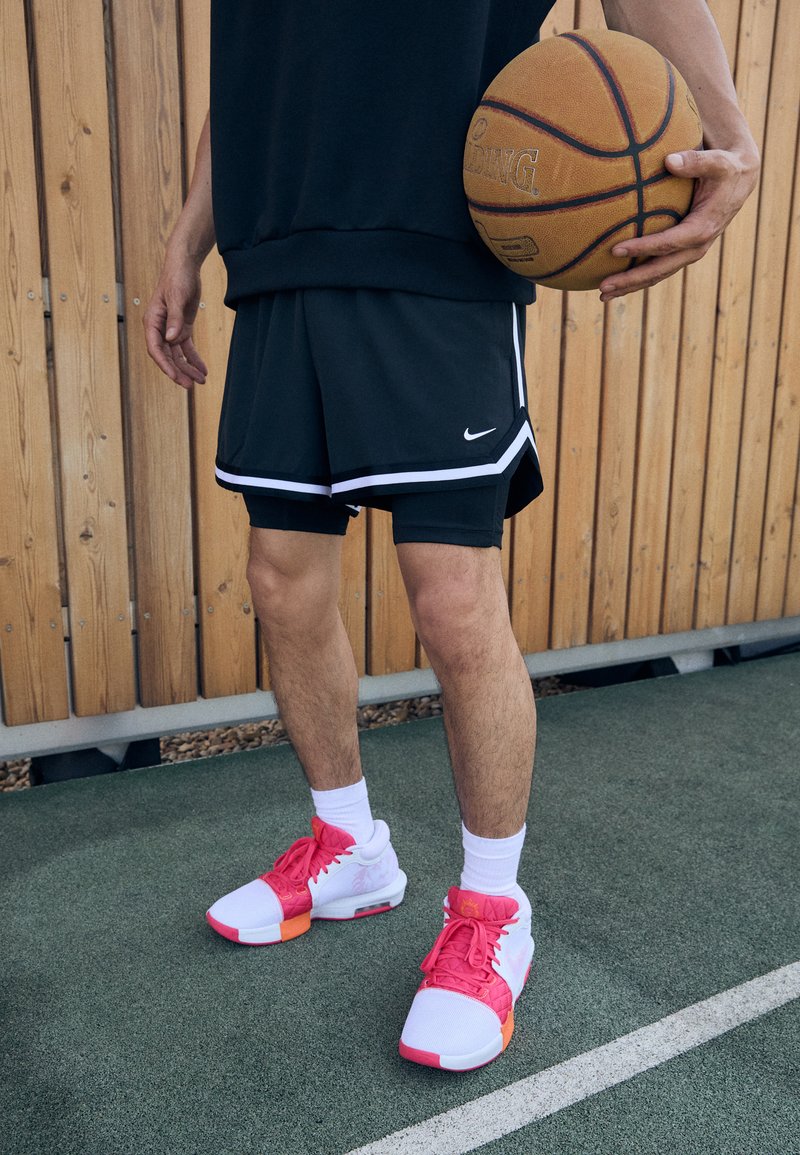 Basketball player in black shorts with white accents, wearing pink and white sneakers, holding a basketball, against a wooden backdrop.