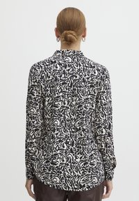 Long-sleeve shirt in white with abstract black pattern, featuring a collar, button-down back, and straight hemline.