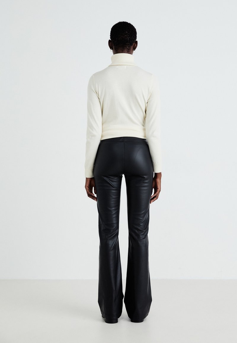 Maglione bianco a collo alto con texture a coste, abbinato a pantaloni neri in eco-pelle a vita alta e a zampa, visti da dietro.