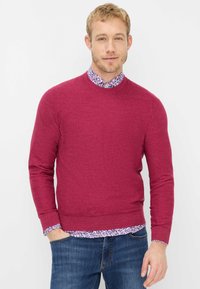 Bordeauxroter Pullover mit Rundhalsausschnitt und gerippten Bündchen, darüber getragen ein Hemd mit Blumenmuster. Kombiniert mit blauen Jeans aus Denim.