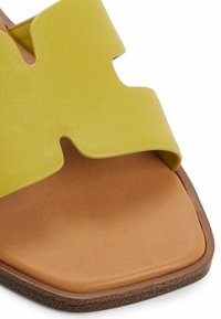 Žluté kožené skládací sandály s širokým, vlnitým páskem a hladkým hnědým stélkou. Čtveratá špička a hnědá texturovaná podrážka.