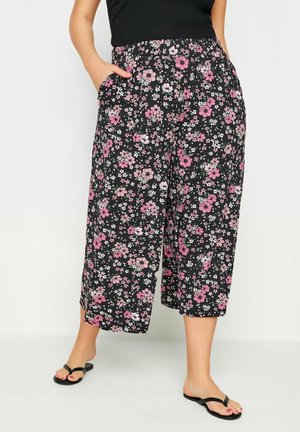 Woman wearing black wide-leg cropped pants with pink and white floral print, black top, and black flip-flops, standing with hands in pockets.