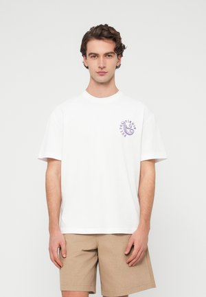 Νεαρός άνδρας στέκεται αντικριστά φορώντας ένα λευκό μπλουζάκι με μοβ λογότυπο χταποδιού στο στήθος και μπεζ σορτς μπροστά σε μια απλή φόντο.