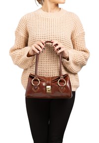 Borsa a mano in pelle marrone con finitura lucida, accessori in oro, manici arrotondati e dettagli decorativi alla base. Maglione lavorato a maglia con motivo sullo sfondo.