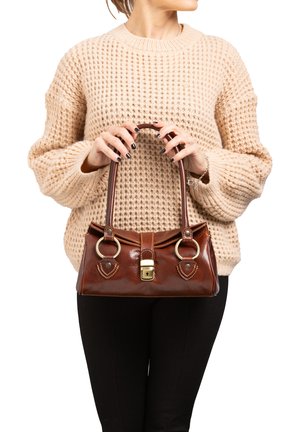 Braune Lederhandtasche mit glänzender Oberfläche, goldenen Beschlägen, runden Griffen und dekorativen Akzenten am Boden. Gemusterter Strickpullover im Hintergrund.