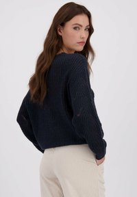 Marineblauwe gebreide trui met een losse pasvorm en een gestructureerd patroon, voorzien van een brede ronde hals en lange mouwen, gecombineerd met lichtbeige geribbelde broek.