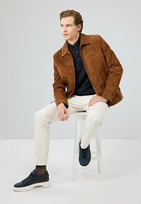 Bruine suède jas met een kraag, zwarte polo, crèmekleurige broek en donkerblauwe sneakers met witte zolen. Model zit op een witte kruk.