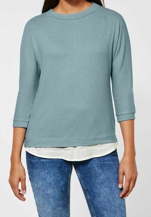 Frau trägt einen hellblauen Langarmpullover über einem weißen Hemd, kombiniert mit blauen Jeans, Torso und Arme sichtbar vor einfarbigem Hintergrund.