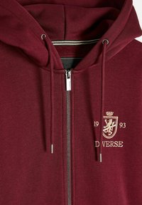 Burgundowa bluza z kapturem zapinana na zamek, wykonana z mieszanki bawełny, z metalowym zamkiem, regulowanym sznurkiem oraz haftowanym herbem z datą i napisem.