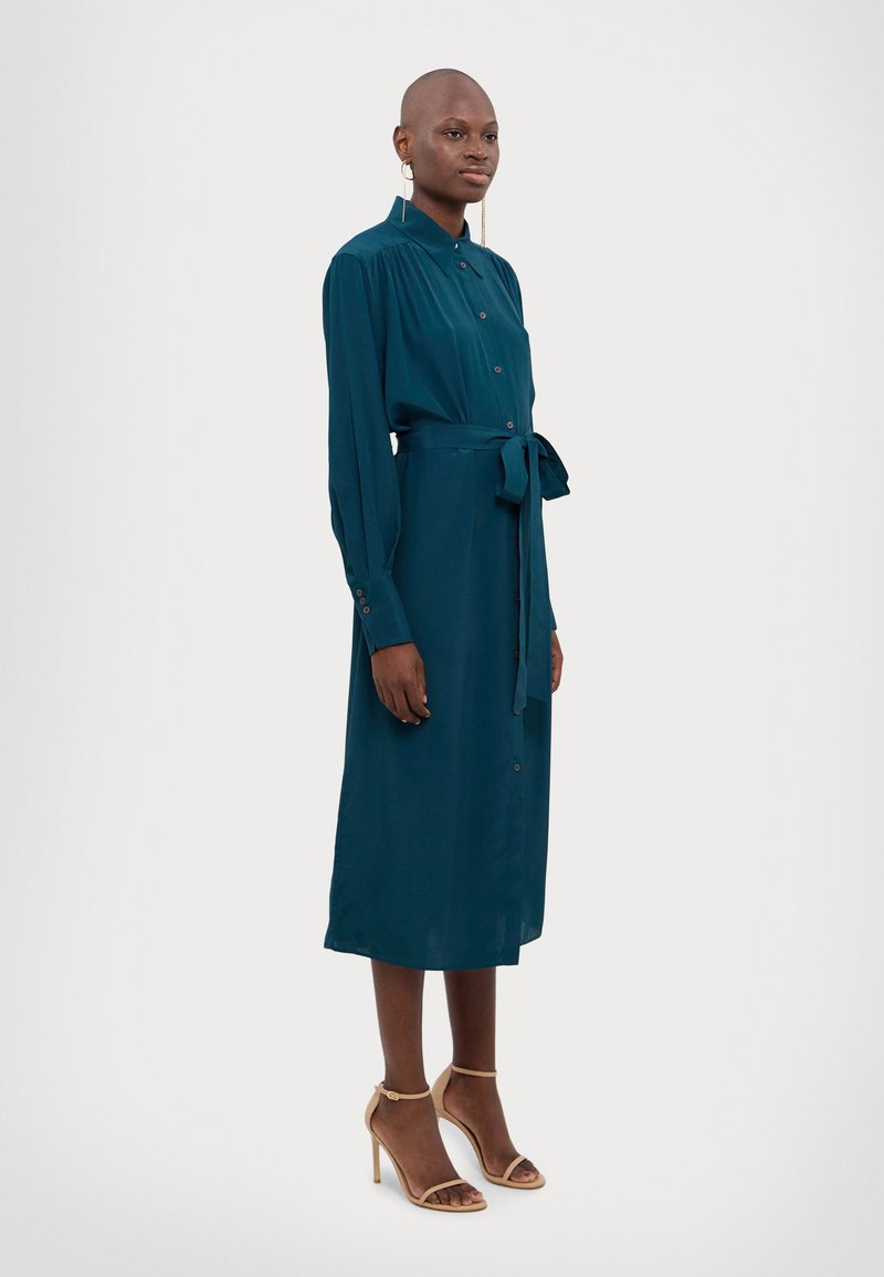 Teal pitkähihaisella napituksella varustettu mekko, jossa on vyötäröllä solmittava vyö. Mekko on valmistettu sileästä kankaasta ja ulottuu polveen. Asusteena on ihonväriset korkokengät.