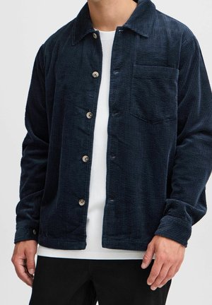 Homme portant une veste boutonnée en velours côtelé bleu foncé sur une chemise blanche et un pantalon noir, debout avec les mains détendues le long du corps.