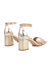 Sandali con tacco dorati in metallo, con tacco a blocco, cinturino alla caviglia, punta aperta e texture liscia. Presentano una silhouette semplice e moderna.